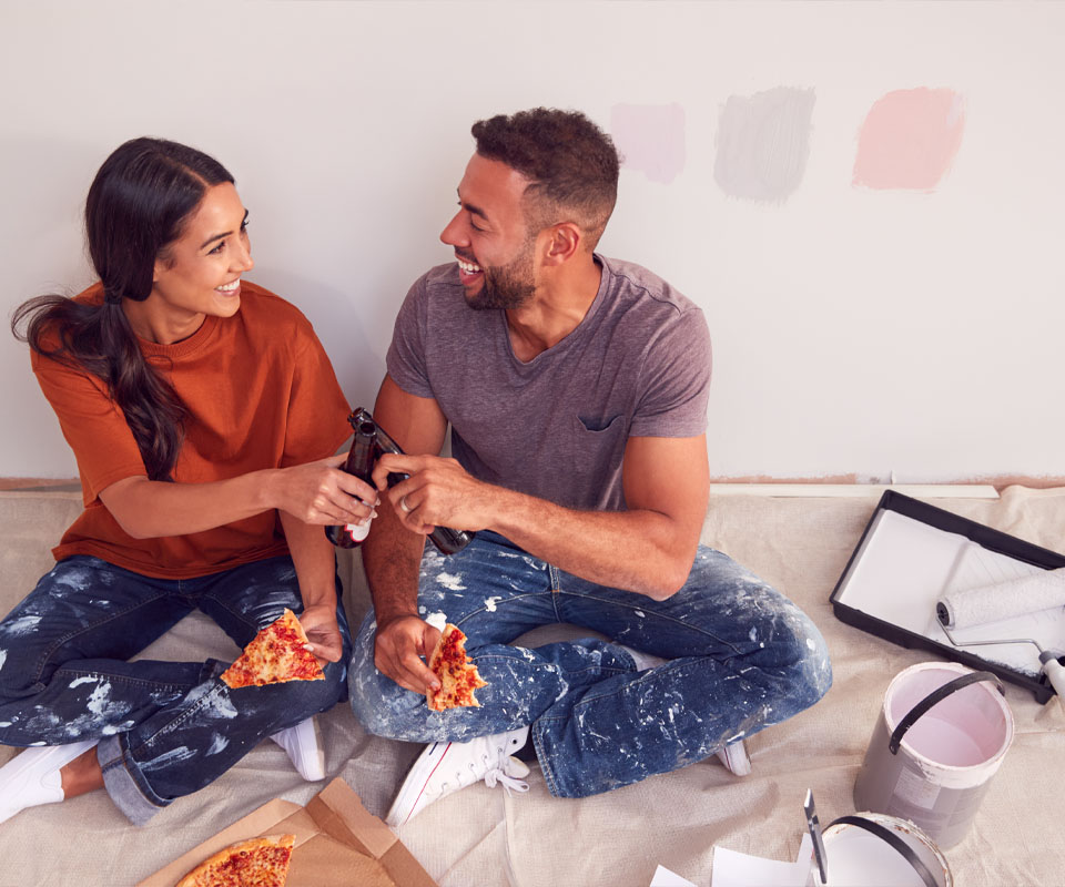 Image showing a happy young couple sat on the floor eating pizza and toasting bottles of beer in front of the wall they are painting