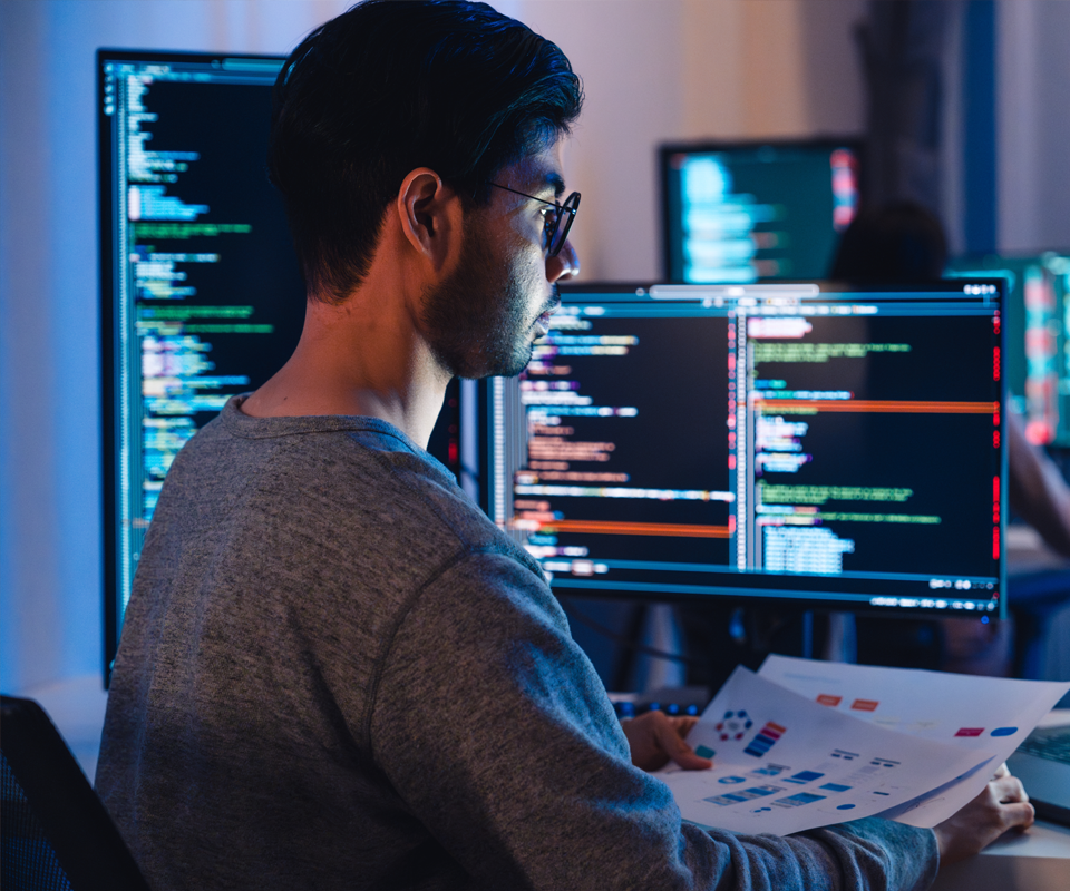 Image showing software developer working on multiple screens displaying code and application diagrams at night in modern office